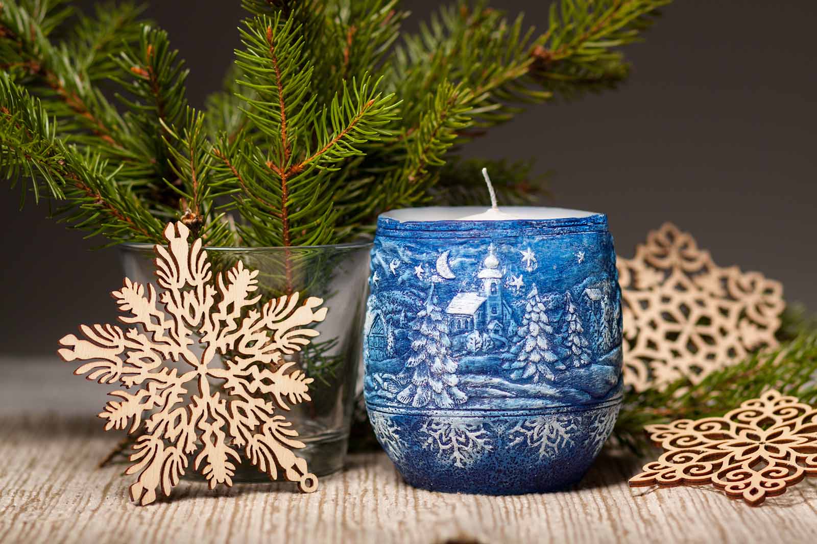 Blue Christmas themed scented candle beside glass vase with pine leaves and snowflake decorations 